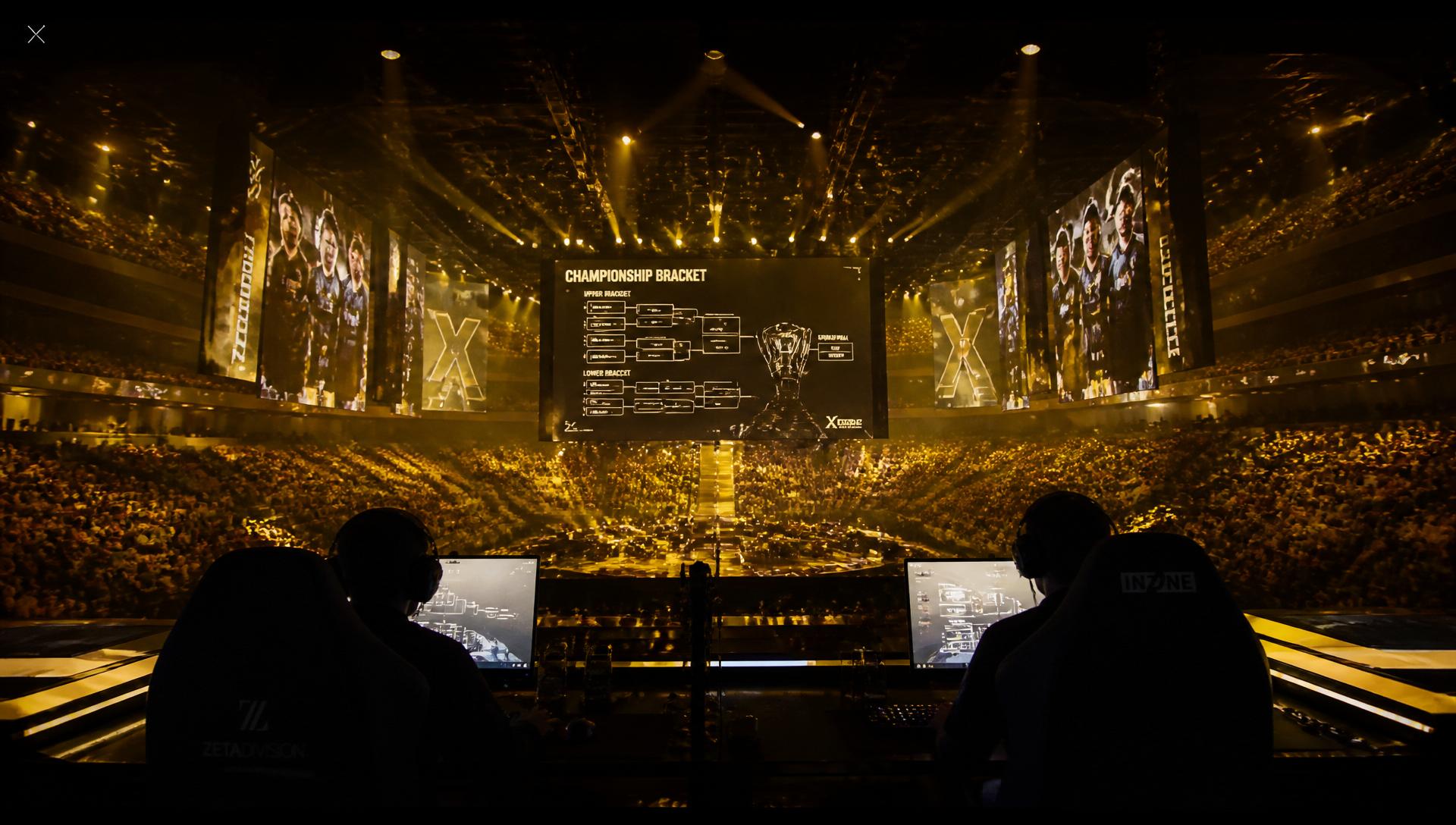 Sold-out esports arena bathed in gold stage lights, silhouetted casters at the desk with a championship bracket on the giant LED wall